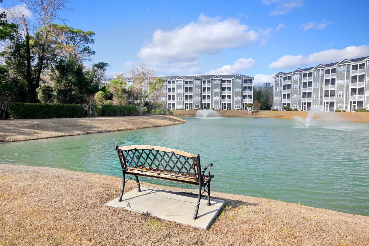 A serene pond with fountains, a comfortable bench, and apartment buildings in the background. The setting focuses on relaxing, reading, meditating, and enjoying nature in a peaceful waterside environment.