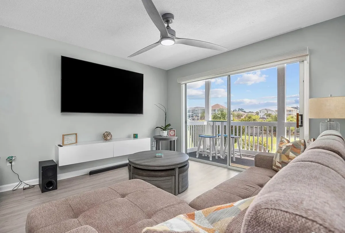 A sunlit living room with a large window offering stunning views of the Intracoastal Waterway, a plush sectional sofa perfect for gathering, a modern ceiling fan above, and a sleek TV mounted on the wall. The space exudes a cozy, welcoming vibe ideal for relaxing family moments while soaking in the coastal scenery.