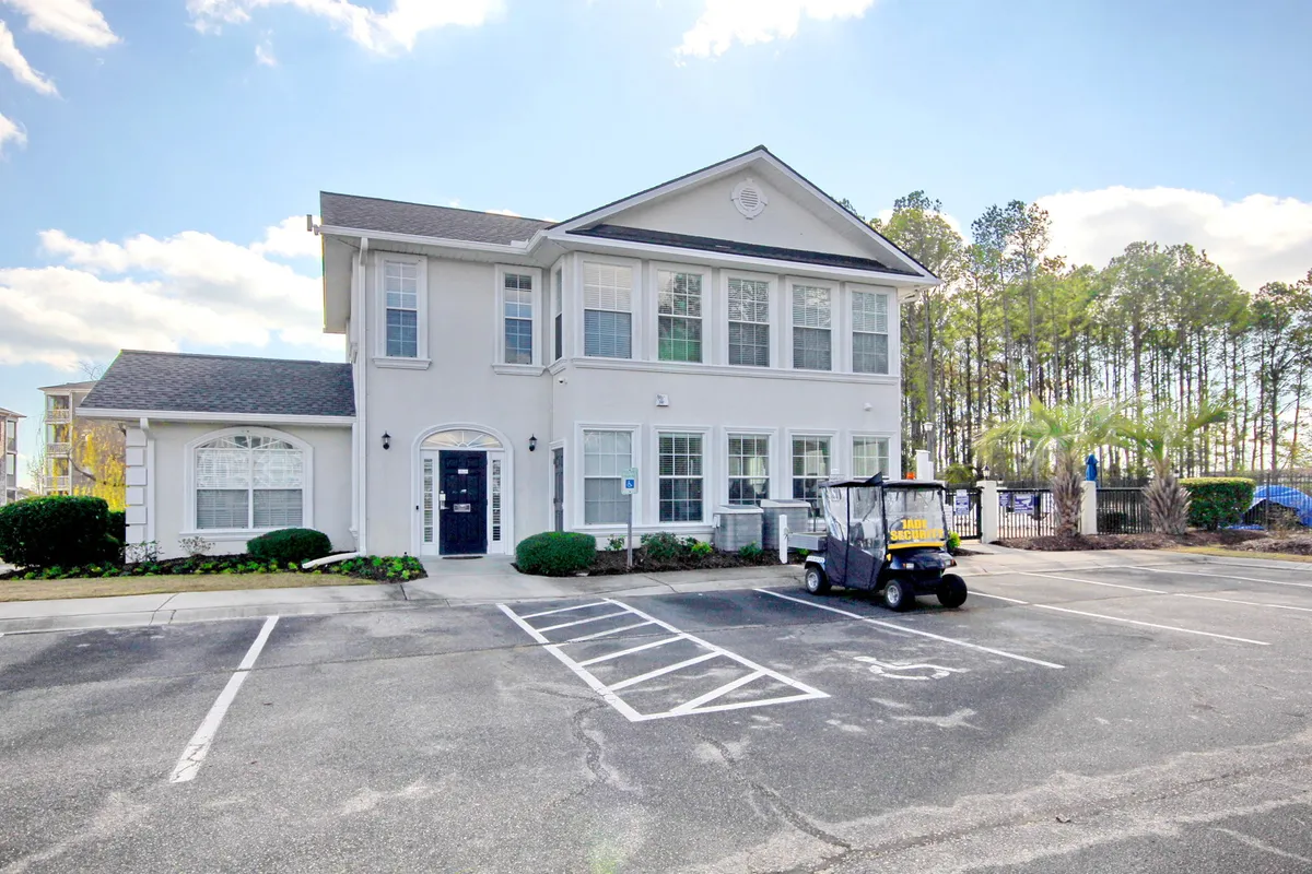 A welcoming two-story clubhouse building set amid landscaped bushes with a golf cart nearby and a paved parking lot. The area suggests a hub for social activity, events, or community recreation.