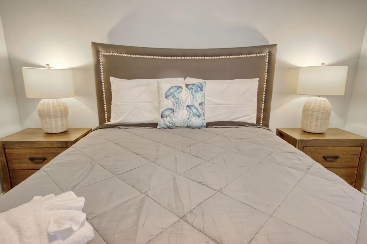 A cozy bedroom with a bed featuring a decorative headboard and a jellyfish-themed pillow, complemented by tasteful nightstands and soft lighting, evoking a sense of relaxation and coastal charm.