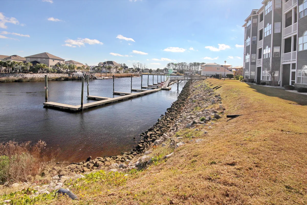 A peaceful dock extends over calm water, bordered by a grassy area with the building facade in view. The scene is tranquil, with sunset reflections glimmering on the water, suggesting a perfect place for relaxation, strolls, or quiet time by the marina.