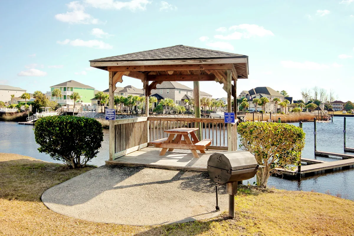 A wooden gazebo with a picnic table and grill overlooks a peaceful water view. The space is ideal for family picnics, barbecues, or simply relaxing together while enjoying the sunset.