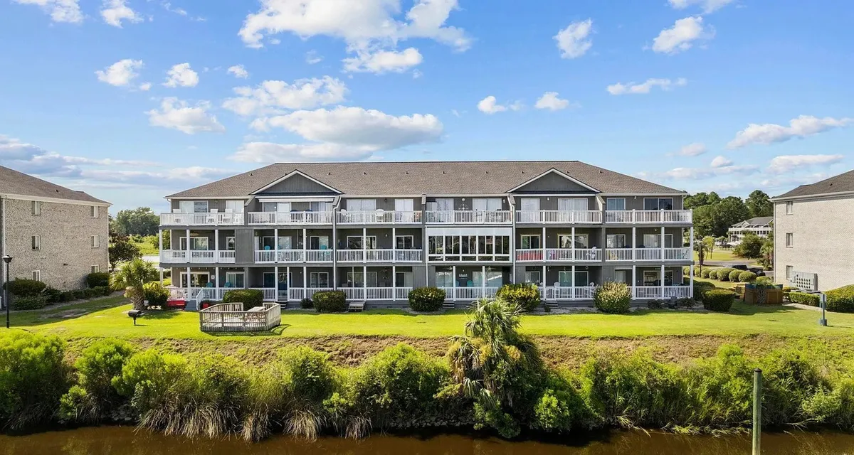A three-story apartment building sits gracefully on a green lawn with a waterfront view. Balconies overlook the water, inviting residents to enjoy peaceful mornings or scenic sunsets. The setting includes well-maintained landscaping and gives a sense of privacy and relaxation by the water.